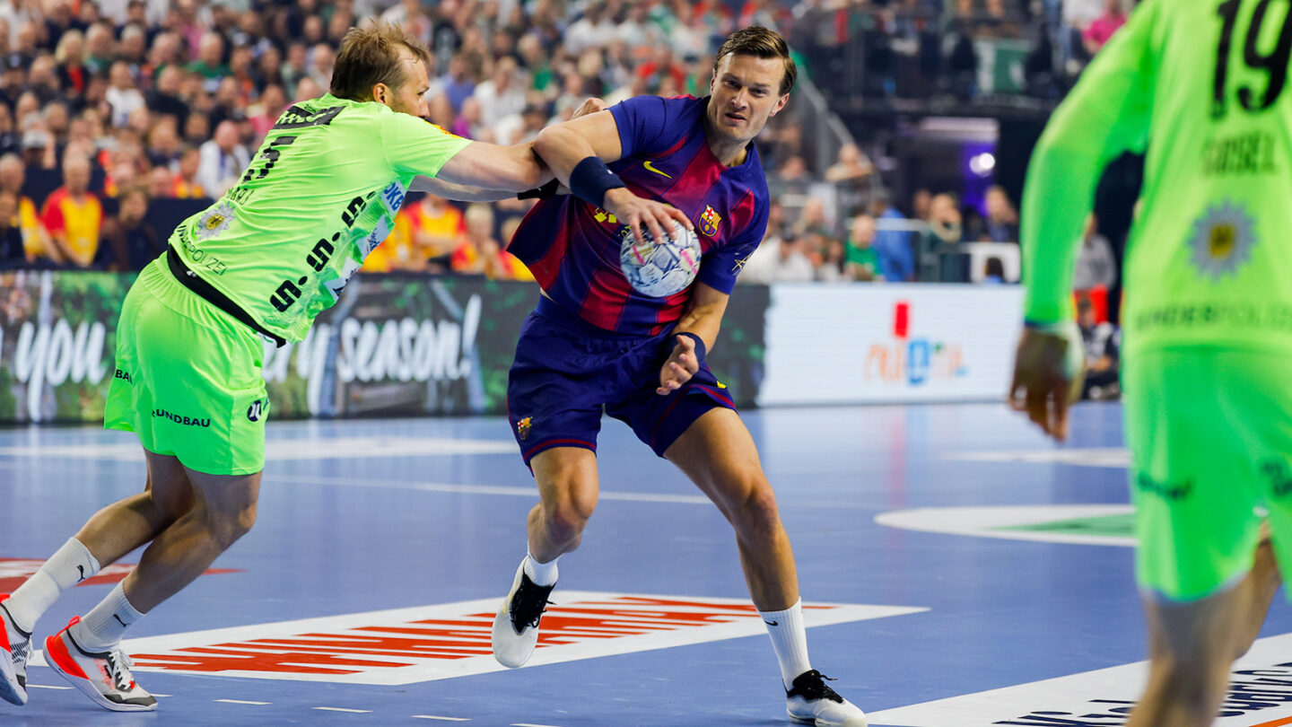A player in a blue jersey holds the ball while being defended by an opponent in a green jersey during an indoor handball game.