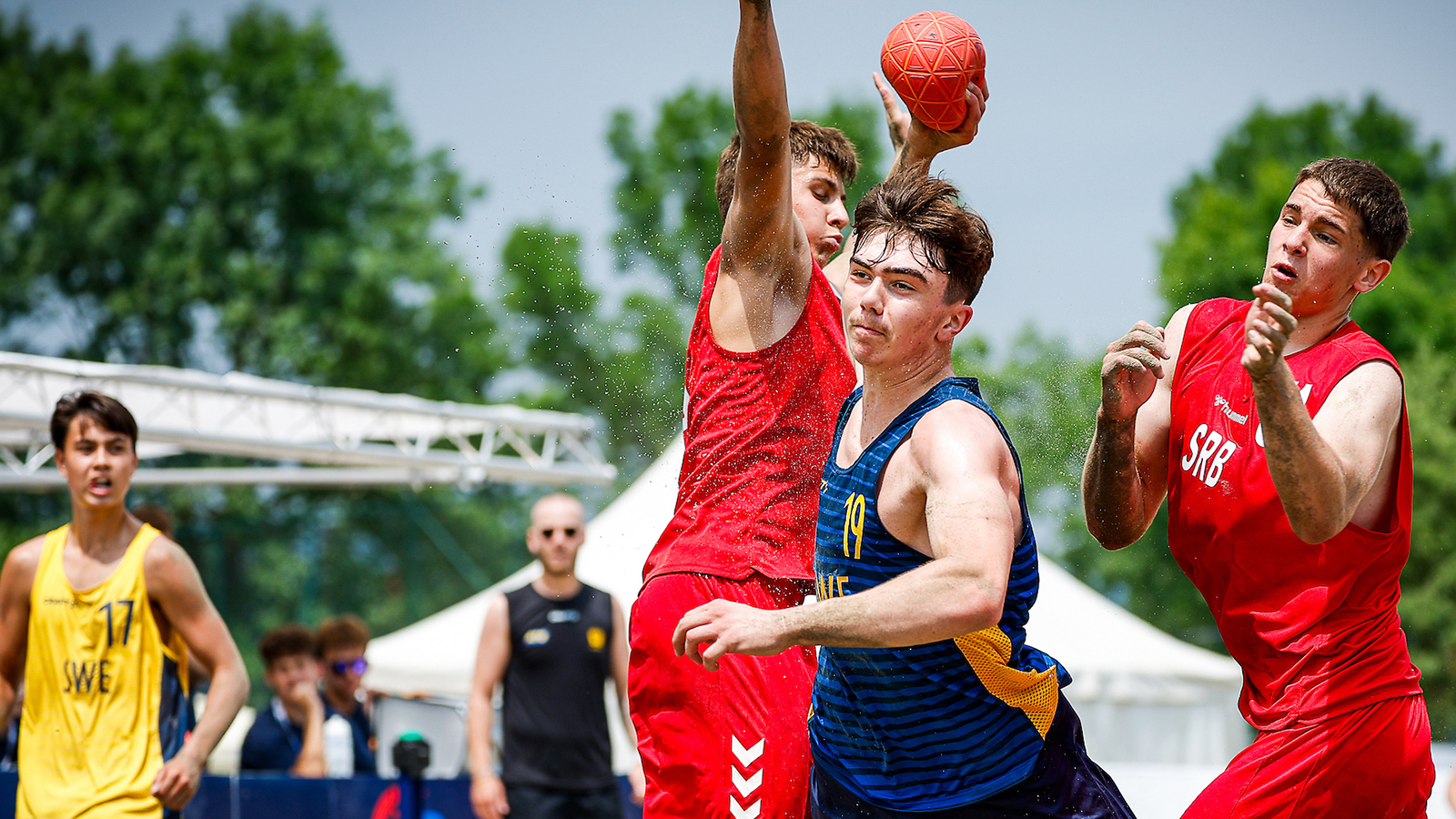 Athletes in red and blue jerseys compete intensely for a ball during a beach handball match on a sandy court.