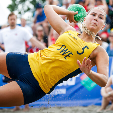 An athlete in a yellow jersey with "SWE 5" dives to throw a green ball during a beach sport match, while another player in red watches nearby.
